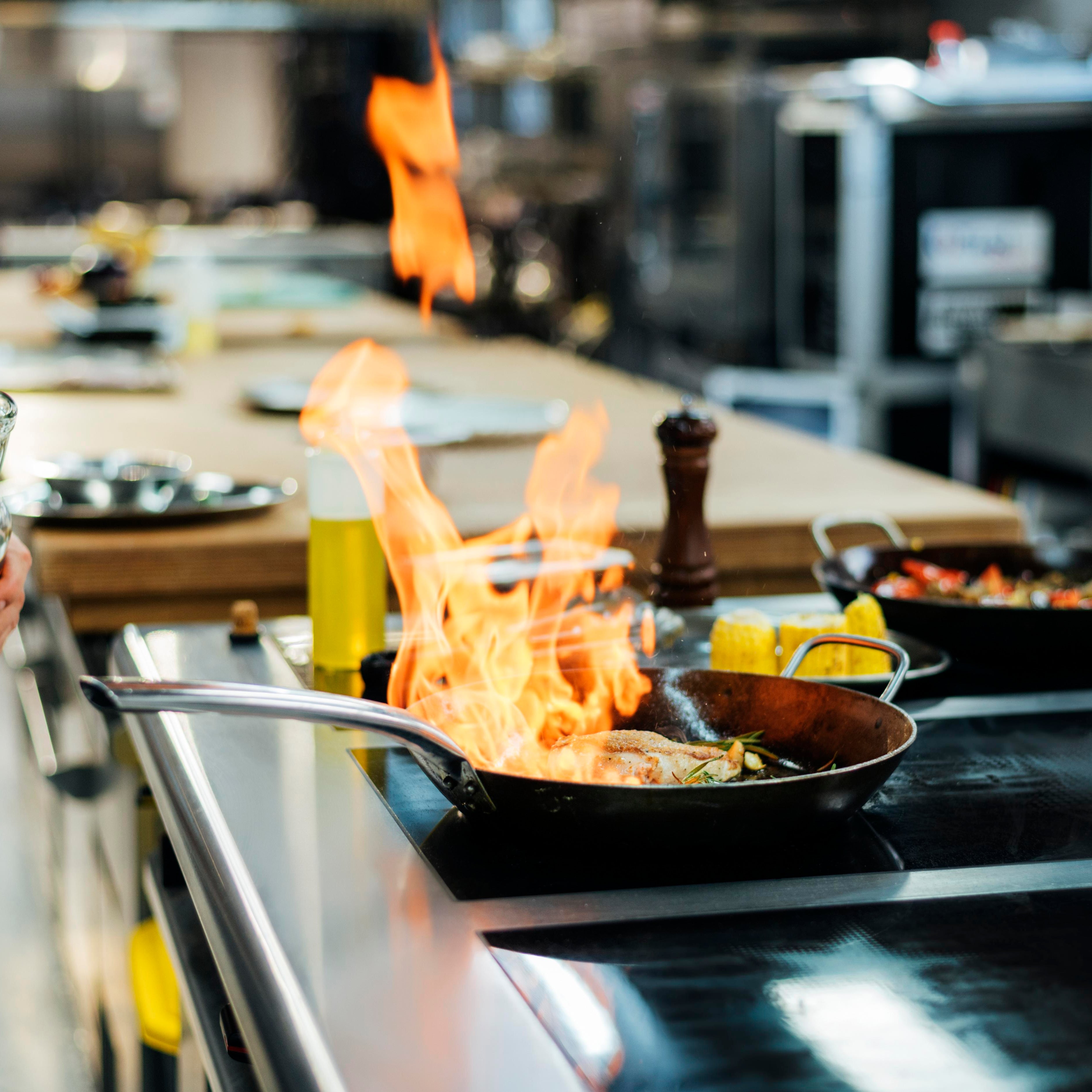 Flaming pan in a professional kitchen with a person holding a bottle.